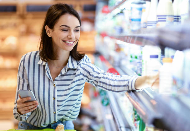 Woman holding smartphone standing in store taking milk Shopping. Young Smiling Woman Holding And Using Phone Buying Food Groceries Standing In Supermarket. Female Customer With Smartphone Taking Milk From The Fridge In Dairy Section At The Shop groceries stock pictures, royalty-free photos & images