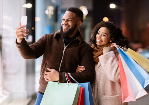 Capturing Bright Moments. Happy excited african american lady and guy taking selfie picture in the city in the evening after shopping, lovers carrying colorful bags with purchases. Relationship