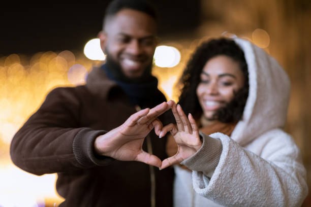 I Love You. Closeup of cheerful positive african american couple making heart shape with fingers walking together in the evening, looking at hands, selective focus, blurred background. Valentine's day