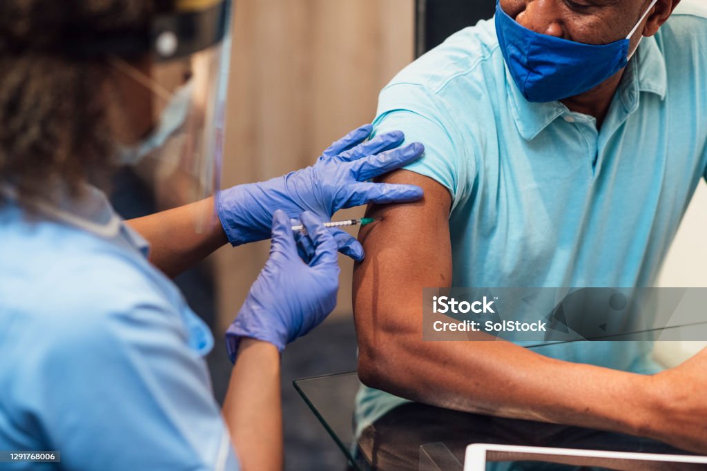 Administering COVID-19 Vaccine A mixed race female nurse wearing a protective face shield, surgical mask and protective gloves administering the COVID-19 vaccine to a senior black man in his home. COVID-19 Vaccine Stock Photo