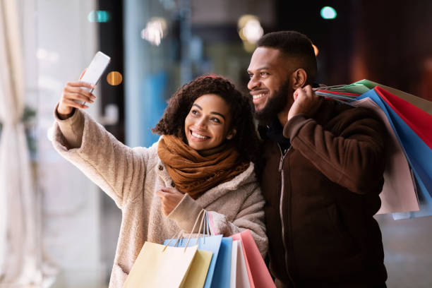 Couple Selfie. Portrait of happy casual african american woman and man taking picture on the street near mall, smiling at camera, holding colorful shopping bags with purchases. Family Photo