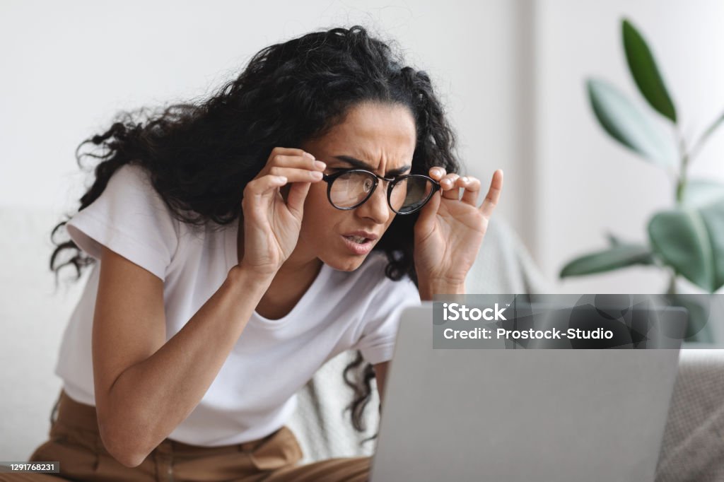 Young woman with bad eyesight using laptop, wearing glasses Young woman freelancer with bad eyesight using laptop, using glasses, home interior, copy space. Curly lady holding her glasses and squinting, looking at laptop screen, having vision troubles Eyesight Stock Photo