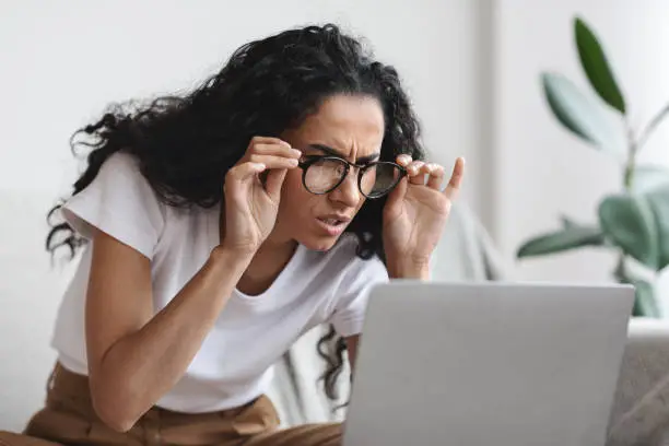 Young woman freelancer with bad eyesight using laptop, using glasses, home interior, copy space. Curly lady holding her glasses and squinting, looking at laptop screen, having vision troubles