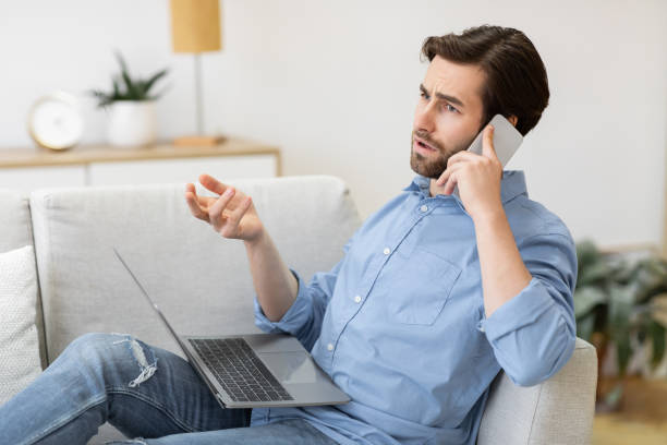 istock Discontented Guy Talking On Cellphone Sitting With Laptop At Home 1291768355