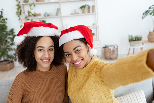 Selfie for New Year and Christmas with friend at home. Happy african american and mixed race teens women in sweaters and santa claus hats making photo in interior of cozy living room, free space