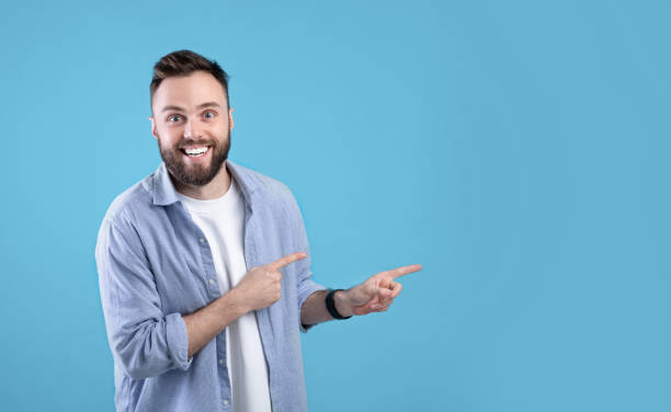 istock Excited young bearded guy smiling and pointing aside at empty space over blue studio background, panorama 1291769828