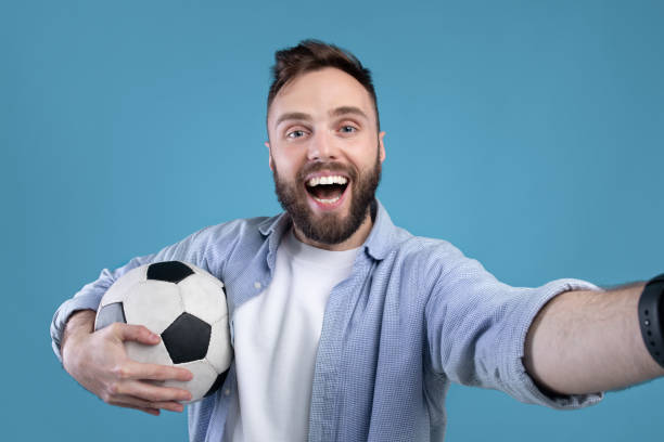 istock Happy soccer fan with ball taking selfie on mobile device over blue studio background 1291769992