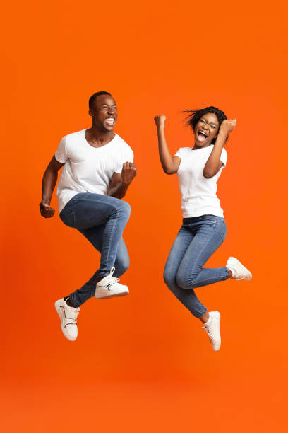Emotional african american couple winners clenching fists and jumping up Emotional african american young couple winners clenching fists and screaming, jumping up over orange studio background. Happy black man and woman celebrating success, full length photo raised fist photos stock pictures, royalty-free photos & images