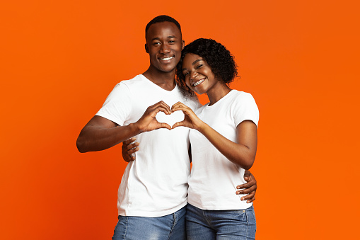 Smiling african american lovers young man and woman cuddling and making together heart shape gesture with their hands as a sign of their love, orange studio background, copy space. Valentine concept