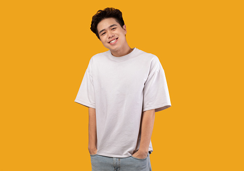 Positive Male Model. Portrait of smiling asian guy wearing blank white t-shirt for design print mockup, standing isolated over orange studio background. Cheerful casual man looking at camera