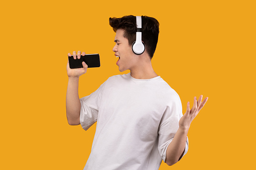 Having Fun. Portrait of overjoyed asian guy singing song and using smart phone as microphone, wearing wireless headset. Carefree young man dancing isolated over orange studio background