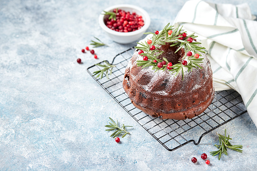 Christmas homebaked dark chocolate bundt cake decorated with powdered sugar and fresh cranberries on metal wire rackin light blue background