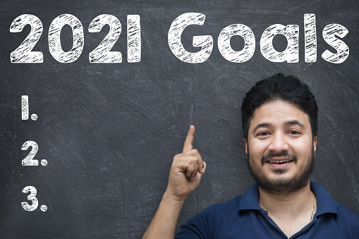 Handsome Indian/Asian young man pointing up while standing against blackboard with chalk drawing of  new year 2021 goals