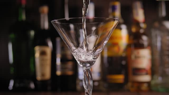 Martini being poured into a glass in a bar with blurred bottles of alcohol.