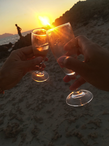 The party is on the beach at sunset. Two hands with glasses and a silhouette of a man in the background near the sea