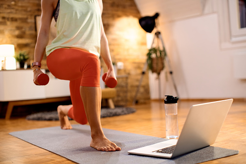 Unrecognizable sportswoman having online training class and practicing lunges while holding hand weights at home.