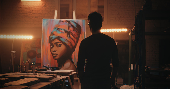Back view of anonymous man admiring portrait of black woman then walking closer to artwork and applying finishing strokes during work in illuminated workshop