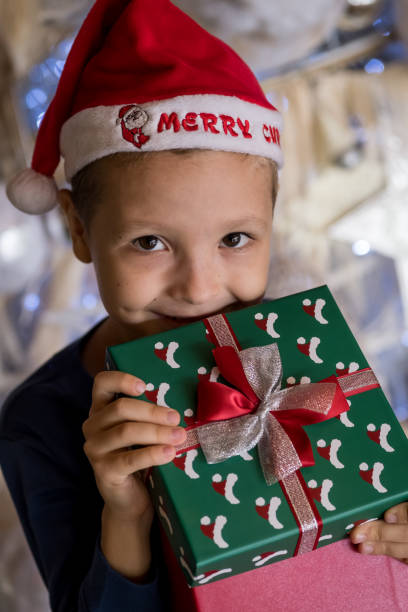 happy little boy wearing santa hat, holding present gift box near christmas tree and looking, peeking inside. merry christmas and happy holidays - christmas child little boys peeking imagens e fotografias de stock