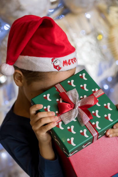 happy little boy wearing santa hat, holding present gift box near christmas tree and looking, peeking inside. merry christmas and happy holidays - christmas child little boys peeking imagens e fotografias de stock