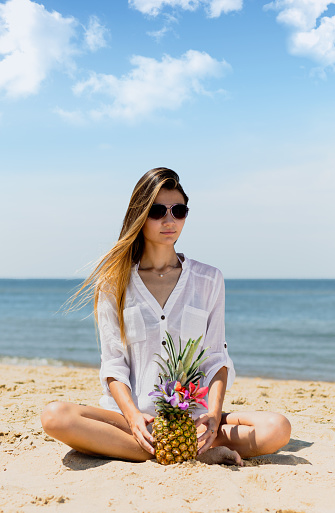 Young woman sitting on beach wearing sunglasses with pineapple