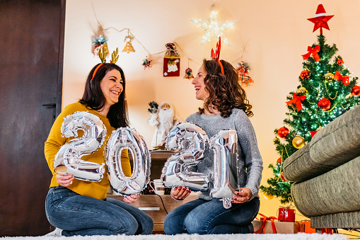Young beautiful female friends celebrating New Year's eve together in beautiful decorated home. They are holding balloon numbers 2021.