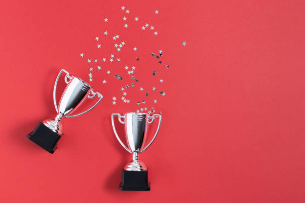 istock Winner or champion silver trophy cups with confetti on red background top view. 1291825216