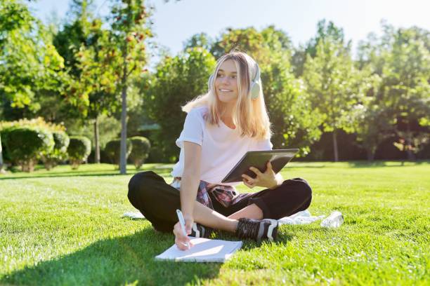 dziewczyna nastolatek student w słuchawkach siedzi w parku na trawie z cyfrowym tabletem piśmie w notatniku - teenagers only lake summer sunlight zdjęcia i obrazy z banku zdjęć