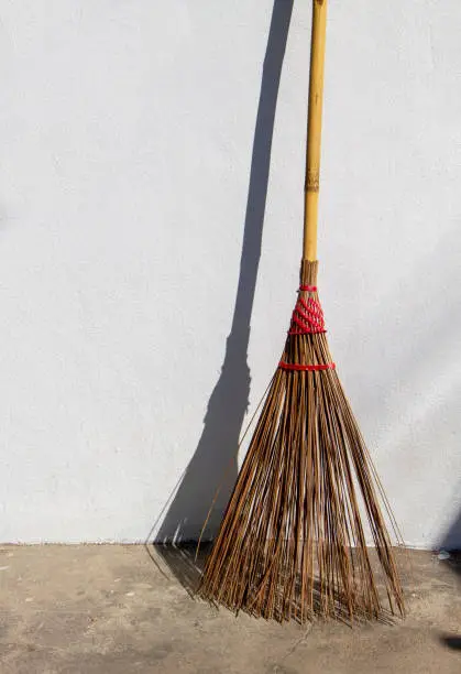 A broom made of coconut palm leaf stalk and bamboo handle lean on a white concrete wall under the afternoon sunlight.