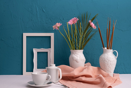 photo frames, a cup with coffee and a teapot and two vases with gerbera flowers and reed cones on a blue background on the table