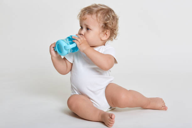 istock Little baby drinking water from blue bottle, looking aside while sitting on floor, posing barefoot and dresses bodysuit, cute infant kid having wavy blond hair. 1291907980