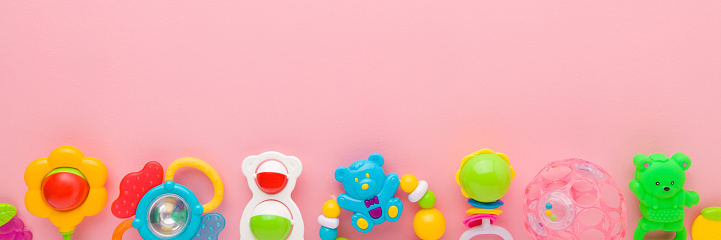 Different colorful rattle toys for baby on light pink table background. Pastel color. Closeup. Empty place for text. Wide banner. Top down view.