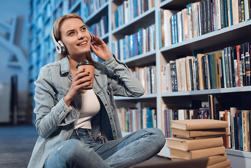 Beautiful girl looks away while sitting in a library with coffee. Girl sitting in a library listening to music and drinking coffee.