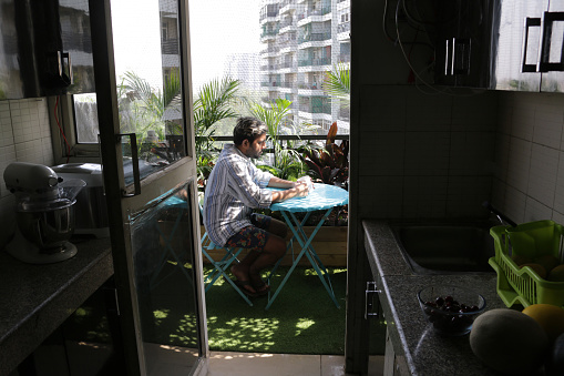 Stock photo of man sat at blue circular table on balcony drinking a mug of hot morning coffee in sunshine with tropical jungle garden in the background, view from gallery kitchen doorway concept stock photo.