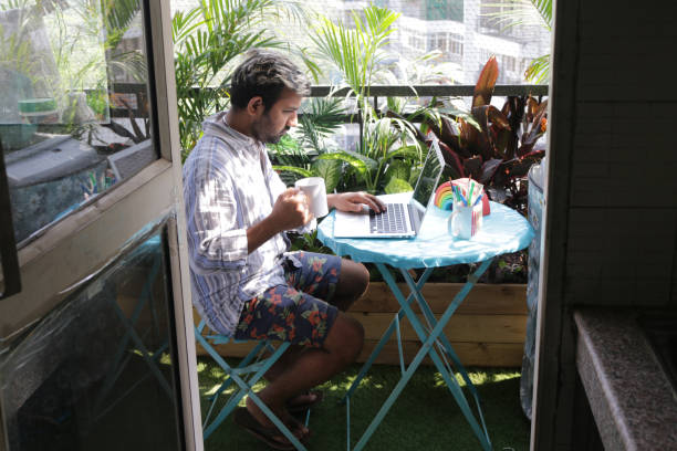 Stock photo of man sat at blue circular table on balcony patio working on a laptop in sunshine, drinking hot coffee with tropical palm jungle garden in the background overlooking the apartment tower block, view from kitchen doorway, Coronavirus new normal working from home concept stock photo. imagen del hombre se sentó en la mesa circular azul en el patio del balcón trabajando en un ordenador portátil en el sol, bebiendo café caliente, fondo del jardín de la selva de palma tropical con vistas al bloque de la torre del apartamento, vista de