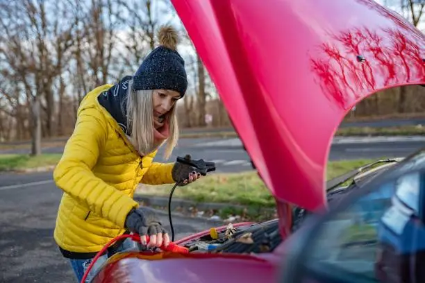 Photo of Beautiful blonde woman in yellow jacket trying to start her broken car with jumper cables.