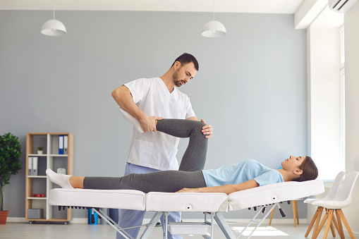 Professional physiotherapist helping woman with knee pain. Osteopathy specialist examining young patient's leg. Female athlete lying on hospital table doing medical exercise with licensed chiropractor