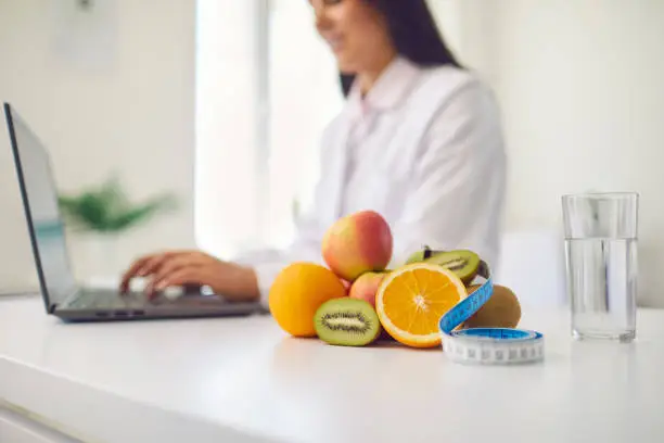 Close-up of fresh apples, kiwis and oranges with measuring tape and glass of clean water on office desk against blurred dietitian with laptop giving online weight loss and healthy diet consultation