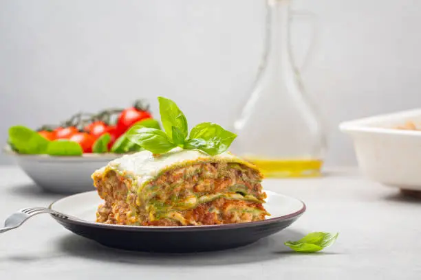 Homemade oven-baked green lasagna - Lasagne alla bolognese with spinach in the dough, ragu - meat sauce, bechamel and parmesan cheese. Italian food. Fresh spinach and tomatoes, olive oil on background. Horizontal image.