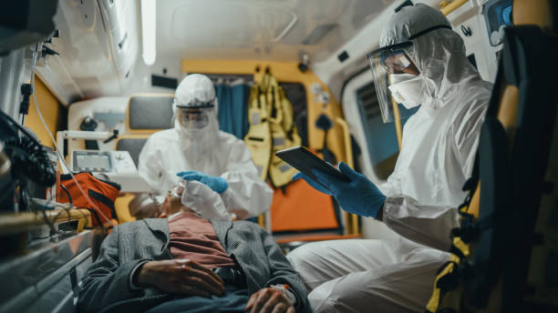 paramédic dans coveralls utilisant l’ordinateur de tablette pour vérifier le diagnostic pour le patient blessé sur le chemin de l’hôpital. les aides médicaux d’urgence mettent un masque de ventilation dans une ambulance. - véhicule de service durgence photos et images de collection