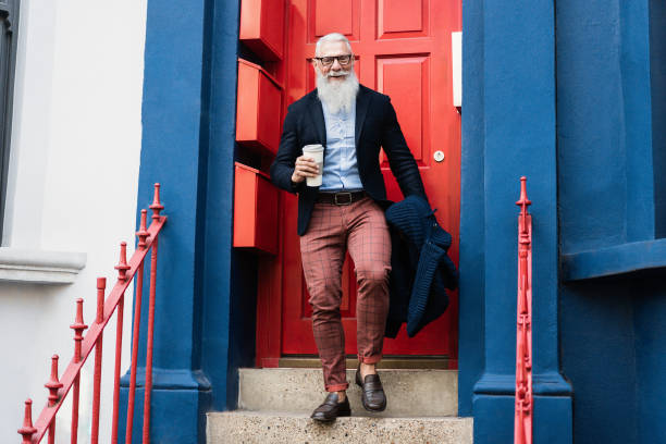 Hipster senior man drinking coffee while walking out with home door in background - Focus on face