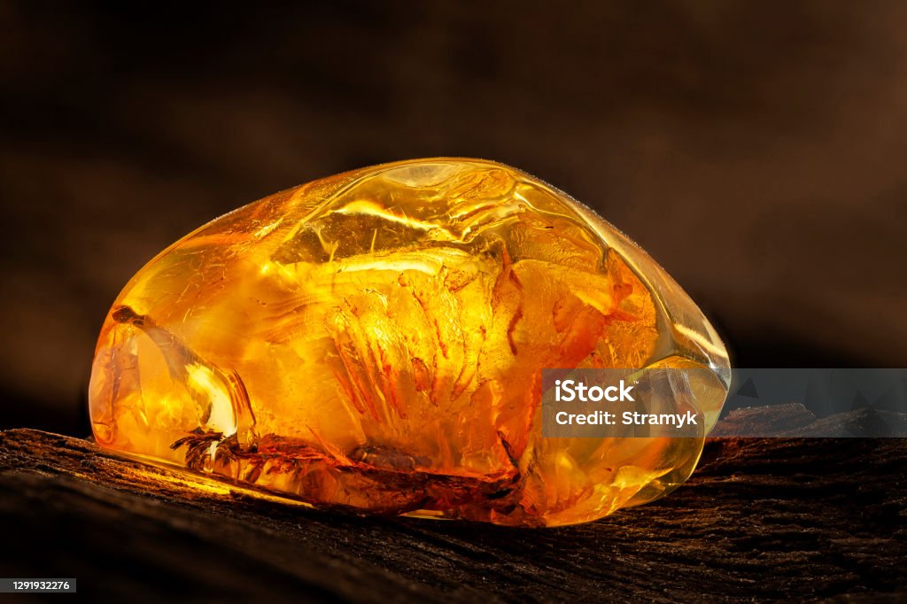 Beauty of natural raw amber. A piece of yellow transparent natural amber on piece of stoned wood A piece of yellow transparent natural amber, classification color honey, has cracks and imprints on its surface and contains pieces of leaf. Placed on dark stoned wood texture. Amber Stock Photo