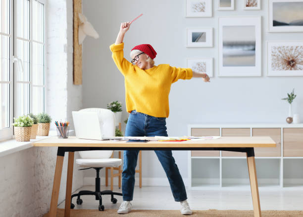 cheerful elderly woman freelancer creative designer in a red hat having fun and dancing in workplace cheerful elderly woman freelancer creative designer in a red hat having fun and dancing in the workplace career vitality stock pictures, royalty-free photos & images
