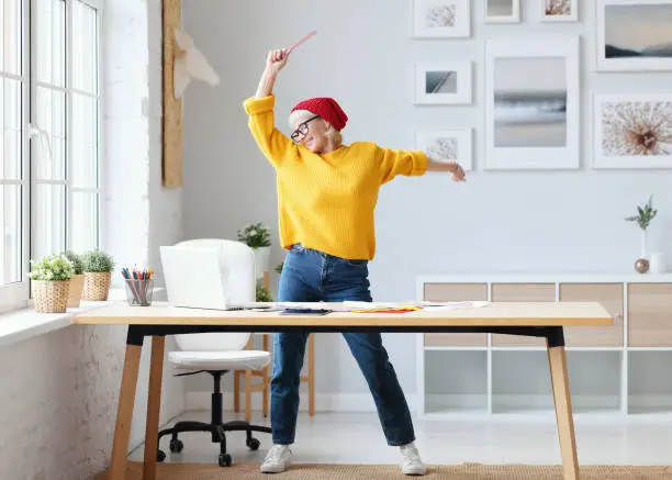 Photo of cheerful elderly woman freelancer creative designer in a red hat having fun and dancing in workplace