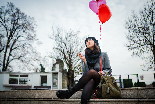 Young attractive woman sitting and smoking with heart - shaped balloons in her hand. Being late for a date / love disappointment / lonely on a Saint valentine's day concept.