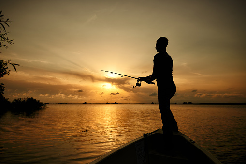 Silhouette fisherman with hat fishing on river bank at sunset time, lifestyle concept