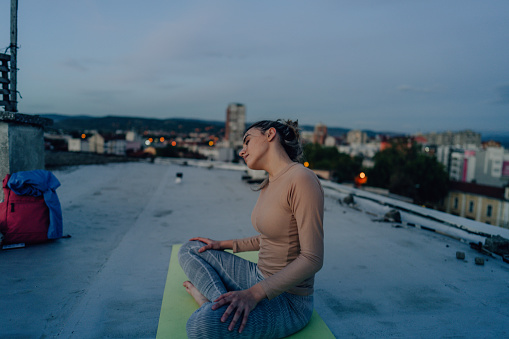 Photo of a young woman, practicing yoga and pilates, to stay healthy and calm. She is performing the exercise on the rooftop of her building, to avoid getting sick from coronavirus.