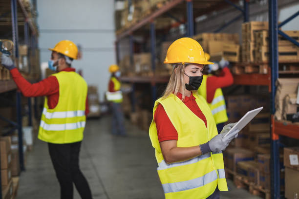 istock Young woman at work in warehouse with digital tablet scanner while wearing protective face mask for coronavirus - People working staff in background 1291958254