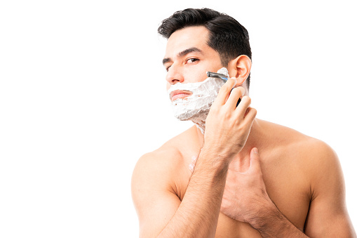 Close-up of a young hispanic male shaving is beard with razor on white background