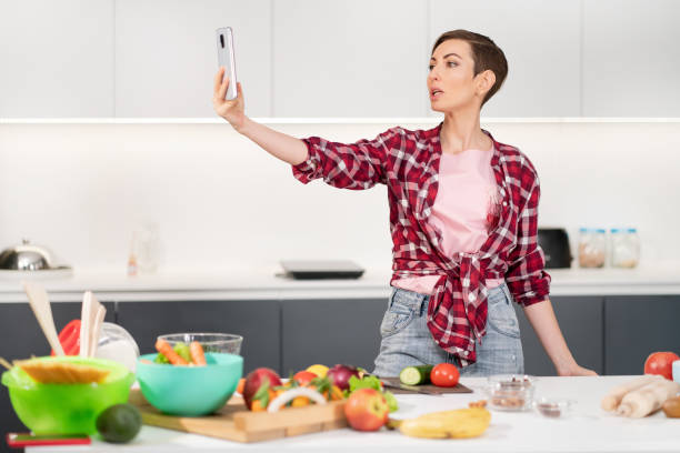istock Pretty woman taking selfie or making a video call using her smartphone holding it in outstretched arm while cooking fresh salad wearing a plaid shirt with a bob hair style 1291962685