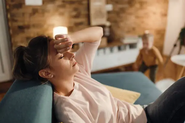 Exhausted mother relaxing with eyes closed on the sofa while her son is playing in the background at home.